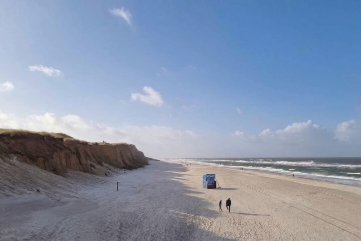 Weiter, fast leerer Sylter Strand am Duenenkliff, in der Mitte ein einzelner blau-weisser Strandkorb mit einem Holzrechen daneben