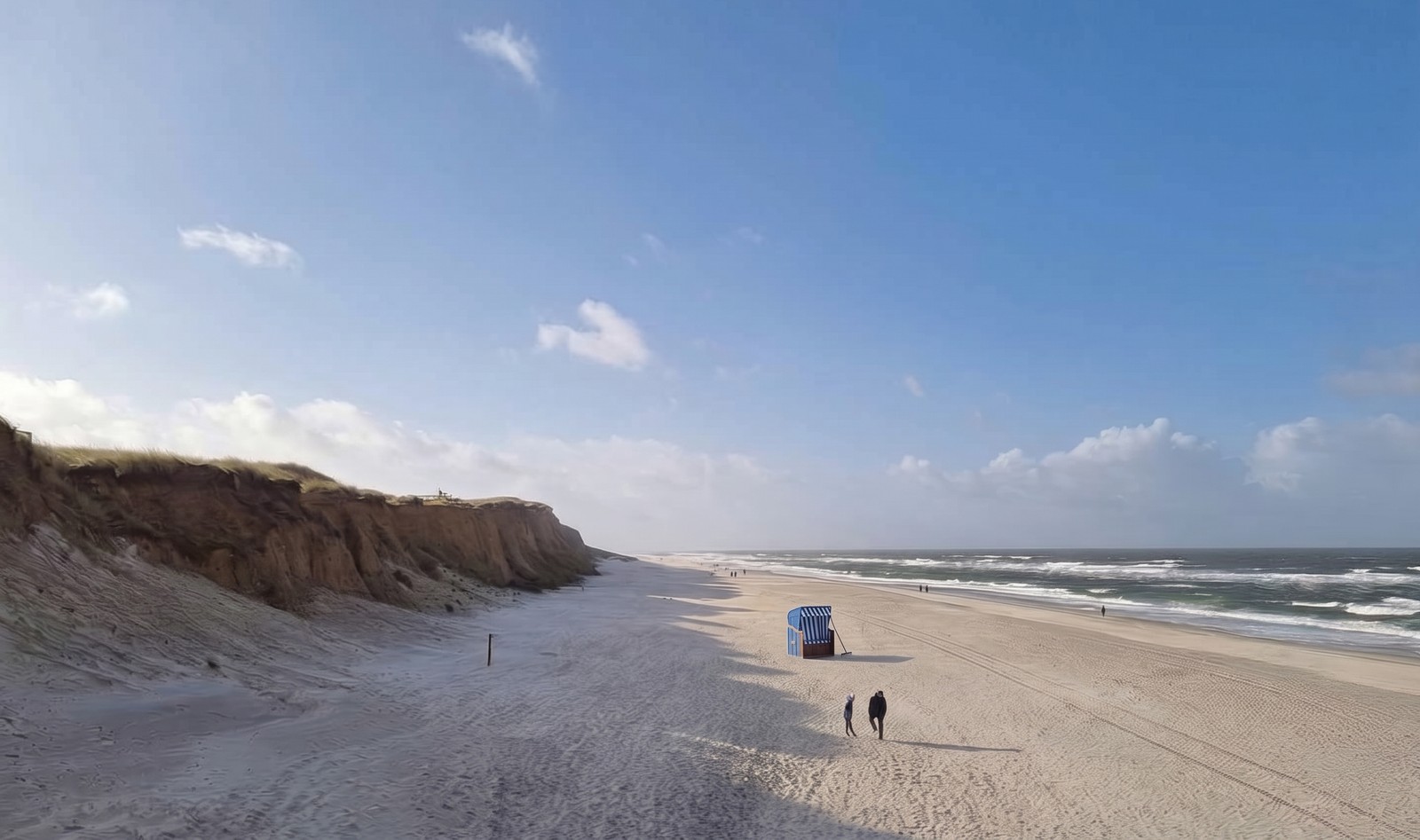 Weiter, fast leerer Sylter Strand am Duenenkliff, in der Mitte ein einzelner blau-weisser Strandkorb mit einem Holzrechen daneben